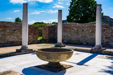 Uzun, beyaz sütunlar Sırbistan 'ın Felix Romuliana kentindeki antik tuğla kalıntılarının arasında duruyor. Arka planda yeşil ağaçlar ve açık mavi bir gökyüzü var.