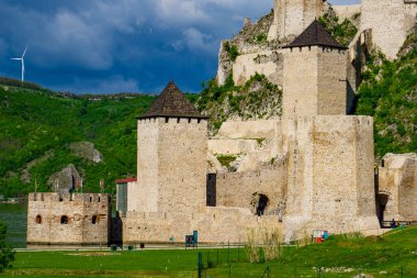 Golubac Kalesi yemyeşil bir zemin ve rüzgâr türbinlerine karşı gururla duruyor, bulutlarla kaplı parlak mavi gökyüzünün altında.