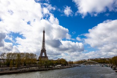 Parlak mavi gökyüzünün altında, ikonik Eyfel Kulesi Seine Nehri 'nin kıyısında duruyor. Etrafı yemyeşil ağaçlar ve Paris' in cazibesini yakalayan yumuşak bulutlarla çevrili.