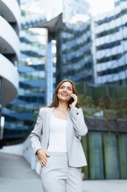 Profesyonel bir kadın neşeli bir telefon görüşmesi yapar, modern binaların önünden geçerken kendine güveni tamdır, aydınlık bir günün sıcaklığında güneşlenir.