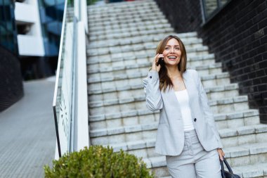 Kendine güvenen takım elbiseli bir kadın şık adımlarla yürüyor, akıllı telefonuna gülüyor, şehir manzarasında profesyonelliğin ve modern hayatın mükemmel bir karışımını sergiliyor.