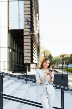 Gri şık bir takım elbiseyle dışarıda gezen, telefonuyla sohbet eden bir kadın. Doğal ışık, güzel yeşilliklerle çevrili güzel gülüşünü güçlendirir.