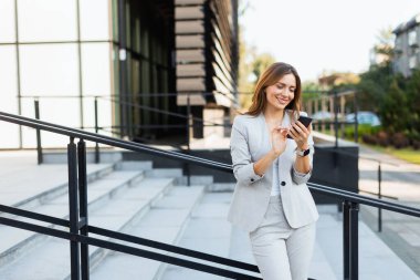 Gri şık bir takım elbiseyle dışarıda gezen, telefonuyla sohbet eden bir kadın. Doğal ışık, güzel yeşilliklerle çevrili güzel gülüşünü güçlendirir.