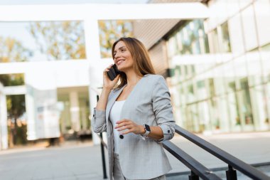 Parlak güneş ışığı, şık bir takım elbise giymiş profesyonel bir kadının yoğun bir iş gününde modern cam bir binanın önünde kendine güvenen bir şekilde konuşmasını vurguluyor.