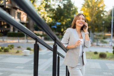 Gri şık bir takım elbiseyle dışarıda gezen, telefonuyla sohbet eden bir kadın. Doğal ışık, güzel yeşilliklerle çevrili güzel gülüşünü güçlendirir.