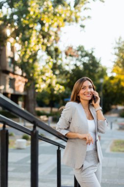 Gri şık bir takım elbiseyle dışarıda gezen, telefonuyla sohbet eden bir kadın. Doğal ışık, güzel yeşilliklerle çevrili güzel gülüşünü güçlendirir.