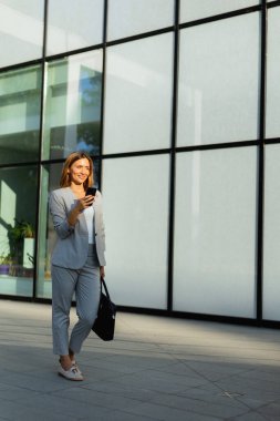 Huzursuz kadın bilerek cam cephe boyunca yürür, telefonuyla meşgul olarak. Güneş ışığı, onun profesyonel görünümünü vurguluyor, zarafet ve modernliğin bir karışımını somutlaştırıyor
