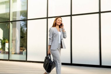 Profesyonel bir kadın zarif cam bir yapıda güvenle dolaşır telefonda konuşurken yüzünde neşeli bir ifade, şık gri bir iş elbisesi giyer.