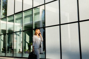 Profesyonel bir kadın zarif cam bir yapıda güvenle dolaşır telefonda konuşurken yüzünde neşeli bir ifade, şık gri bir iş elbisesi giyer.