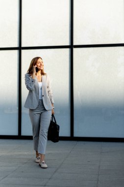 Profesyonel bir kadın zarif cam bir yapıda güvenle dolaşır telefonda konuşurken yüzünde neşeli bir ifade, şık gri bir iş elbisesi giyer.