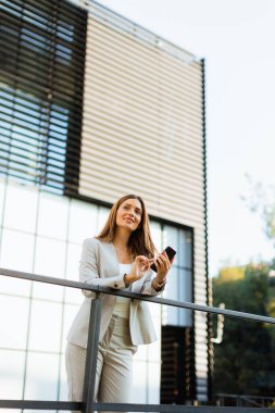 Neşeli iş kadını bir balkonda duruyor, akıllı telefonuyla modern mimarinin arka planına yaslanıyor, sıcak güneş ışığı ve canlı atmosferde sırılsıklam oluyor.