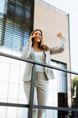 Neşeli iş kadını dışarıda bir anı kutluyor, zarif bir binanın yanındaki korkuluklara yaslanarak neşeli bir telefon görüşmesi yapıyor..
