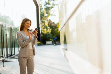 Profesyonel bir kadın rahat yürür, telefonuna odaklanırken gülümser, modern yaşam tarzını arka plandaki doğanın sükunetiyle harmanlar.