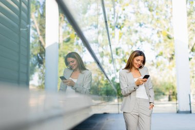 Profesyonel bir kadın rahat yürür, telefonuna odaklanırken gülümser, modern yaşam tarzını arka plandaki doğanın sükunetiyle harmanlar.