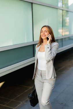 Sıcak bir gülümsemeyle, profesyonel bir kadın, ağaçların arasından süzülen güneş ışığı gibi, zarif bir koridor boyunca güvenli bir şekilde yürür, telefonuyla sohbet eder.