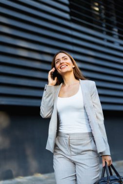 Şık kıyafetli bir iş kadını, parlak bir günde, kendine güveni olan tavırlarını göstererek şık bir mimari zemine karşı telefon konuşmasından zevk alır.