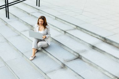 Basamaklarda oturan iş elbiseli bir kadın, güneşli bir günde profesyonelliği dışarıdaki enerjiyle harmanlayarak dizüstü bilgisayarına odaklanıyor.