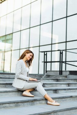 Şık giyinmiş genç bir iş kadını dizüstü bilgisayarının basamaklarında rahat bir şekilde oturuyor. Sıcak güneş ışığı yumuşak gölgeler bırakıyor ve verimliliği canlı bir şehir atmosferiyle harmanlıyor.