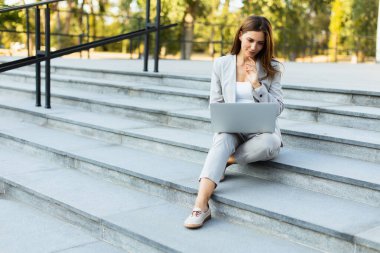 Şık giyinmiş genç bir iş kadını dizüstü bilgisayarının basamaklarında rahat bir şekilde oturuyor. Sıcak güneş ışığı yumuşak gölgeler bırakıyor ve verimliliği canlı bir şehir atmosferiyle harmanlıyor.