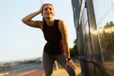 Kadın akşam antrenmanı sırasında durur, güneş batarken kaşlarını silerken, sakin bir yürüyüş yolu ve hafifçe aydınlatılmış bir manzara ile çevrelenir.