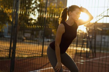 Günbatımının altın parıltısında, odaklanmış bir kadın çitlere yaslanır, enerji veren bir egzersizden sonra nefesini tutar, dayanıklılık ve sebatın vücut bulmuş halidir.