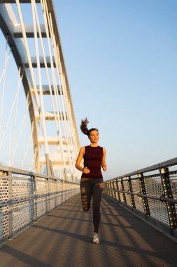 Güneş doğarken, kadın güven içinde köprüden geçiyor, sabah ışığını kucaklıyor ve canlandırıcı koşusu sırasında açık hava serinliyor.