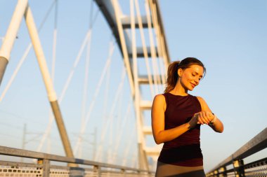 Gün batımının sıcacık parıltısıyla ışıldayan, kendini köprüye adamış bir koşucu, saatini kontrol etmek için bir köprüde duruyor, antrenman rutinindeki azim ve atletizm ruhunu somutlaştırıyor.