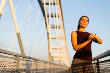 Gün batımının sıcacık parıltısıyla ışıldayan, kendini köprüye adamış bir koşucu, saatini kontrol etmek için bir köprüde duruyor, antrenman rutinindeki azim ve atletizm ruhunu somutlaştırıyor.