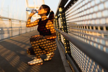 Kararlı bir kadın su içmek için ferahlatıcı bir mola alır, yaya köprüsünde durur. Altın saat güneş ışığı büyüleyici gölgeler yaratır, batışı aydınlatır.