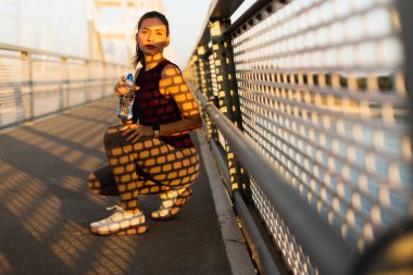 Aydınlık bir günde, bir kadın bir köprüde dinlenir, elinde bir su şişesi tutarken güneş ışığı korkuluklardan süzülür, üzerine karmaşık gölgeler bırakır.