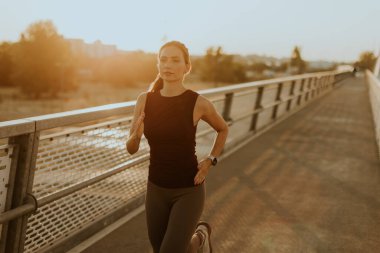 Odaklanmış dişi koşucu zarif bir şekilde parlak bir köprüde hareket eder. Güneşin sıcaklığının tadını çıkararak spor yapma yolunda ilham verici bir ışıltı saçar.