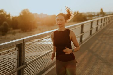 Odaklanmış dişi koşucu zarif bir şekilde parlak bir köprüde hareket eder. Güneşin sıcaklığının tadını çıkararak spor yapma yolunda ilham verici bir ışıltı saçar.