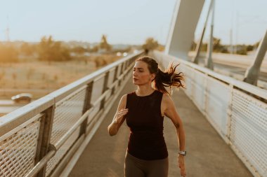 Odaklanmış dişi koşucu zarif bir şekilde parlak bir köprüde hareket eder. Güneşin sıcaklığının tadını çıkararak spor yapma yolunda ilham verici bir ışıltı saçar.