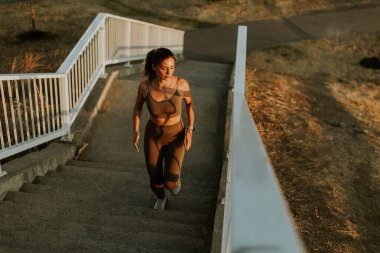 Gün batımının sıcacık parıltısının altında atletik giysiler içindeki bir kadın akşam sporu yolculuğunun somut gücü ve adanmışlığıyla merdivenlerden aşağı iner.