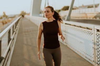 Odaklanmış dişi koşucu zarif bir şekilde parlak bir köprüde hareket eder. Güneşin sıcaklığının tadını çıkararak spor yapma yolunda ilham verici bir ışıltı saçar.