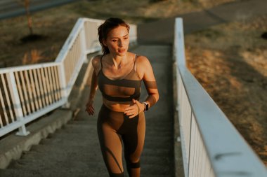 Gün batımının sıcacık parıltısının altında atletik giysiler içindeki bir kadın akşam sporu yolculuğunun somut gücü ve adanmışlığıyla merdivenlerden aşağı iner.