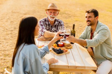 Üç arkadaş, şarap, taze meyve ve neşe ve kahkaha dolu güneşli bir günde, bir üzüm bağında, bir kır masasının etrafında toplanır.