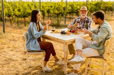Üç arkadaş, şarap, taze meyve ve neşe ve kahkaha dolu güneşli bir günde, bir üzüm bağında, bir kır masasının etrafında toplanır.