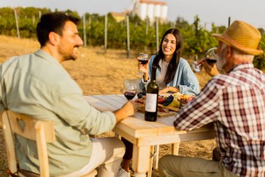 Üç arkadaş, şarap, taze meyve ve neşe ve kahkaha dolu güneşli bir günde, bir üzüm bağında, bir kır masasının etrafında toplanır.
