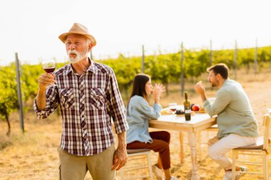 Gün batımının sıcacık parıltıları altında, gülümseyen bir beyefendi, bir bardak kırmızı şarap kaldırır, arkadaşlarıyla çevrili, yemyeşil üzüm bağları arasında keyifli anlar yaşar ve hikayelerini paylaşır.