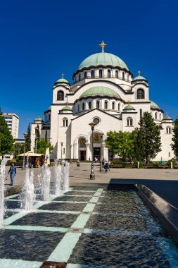 Belgrad, Sırbistan - 29 Nisan 2024: Sırbistan 'ın Belgrad kentindeki Saint Sava Tapınağı, açık mavi gökyüzü ve ikonik beyaz kiliseyi çevreleyen yemyeşil bir yer. Dünyanın en büyük ikinci Ortodoks kilisesi..