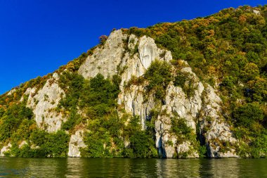 Tuna Nehri 'nin sakin suları, parlak mavi gökyüzünün altındaki görkemli kaya oluşumlarına karşı yemyeşil çimlerin zıt olduğu Djerdap vadisinin yüksek uçurumlarını kucaklıyor.