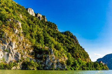 Tuna Nehri 'nin sakin suları, parlak mavi gökyüzünün altındaki görkemli kaya oluşumlarına karşı yemyeşil çimlerin zıt olduğu Djerdap vadisinin yüksek uçurumlarını kucaklıyor.