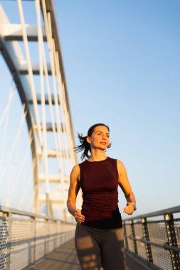 Güneş doğarken, kadın güven içinde köprüden geçiyor, sabah ışığını kucaklıyor ve canlandırıcı koşusu sırasında açık hava serinliyor.