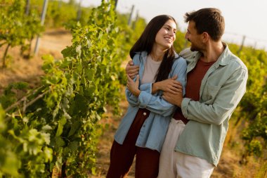 Genç çift yemyeşil sarmaşıklar arasında neşeli bir anı paylaşıyor, sıcak güneşin altında gülüyor ve sarılıyorlar, aşkı ve bir üzüm bağının sükunetini kutluyorlar.