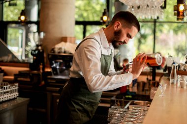 Canlı bir bar ortamında, kendine güvenen bir barmen yeni bir kokteyl hazırlar, uzmanlığını elinde bir şişeyle gösterir, gecenin telaşına hazırdır.