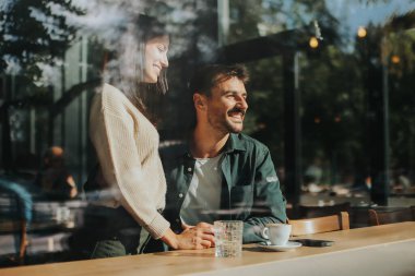 İki arkadaş büyüleyici bir kafede kahkaha ve sıcak içecekleri paylaşıyor, güneşli bir günde bağlantı ve neşeyi kutlayan davetkar bir ortam yaratıyorlar.