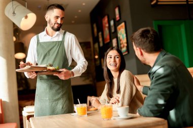 Renkli sanat eserleriyle dolu canlı bir kafede, bir garson, gülen bir çifte taze vejetaryen yemekleri sunar.