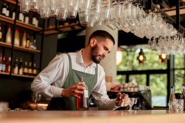 Canlı bir bar ortamında, kendine güvenen bir barmen yeni bir kokteyl hazırlar, uzmanlığını elinde bir şişeyle gösterir, gecenin telaşına hazırdır.