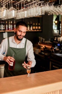 Canlı bir bar ortamında, kendine güvenen bir barmen yeni bir kokteyl hazırlar, uzmanlığını elinde bir şişeyle gösterir, gecenin telaşına hazırdır.